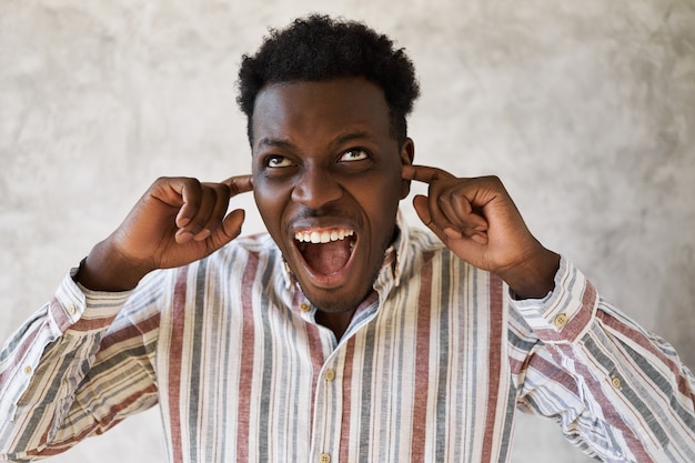 Isolated image of angry stressed young dark skinned male looking up and yelling, shutting ears with fingers, refusing to listen to bad or unpleasant news.