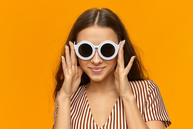 Isolated image of attractive fashionable young woman in striped top trying on round shades in white