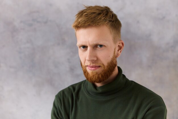 Isolated image of attractive red haired bearded young Caucasian male dressed in elegant stylish clothes staring with intense concentrated look. Human facial expressions and attitude