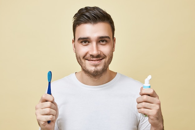 目覚めた直後に歯を磨く、ブラシと歯磨き粉のチューブを保持している剛毛を持つ自信を持って陽気な若いブルネットの男の孤立した画像。衛生、朝のルーチンと歯のホワイトニングの概念