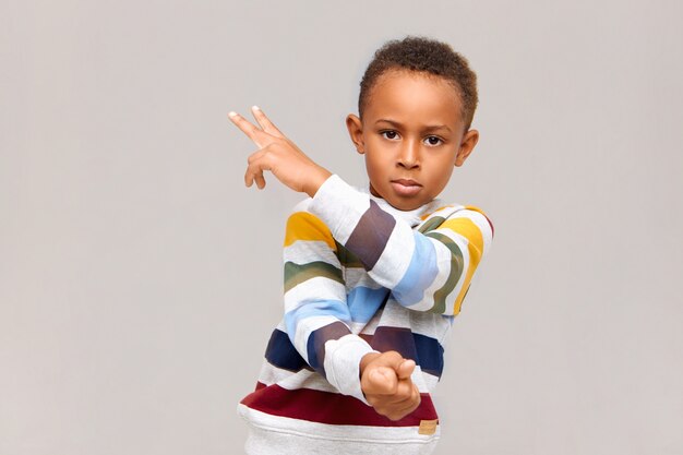Isolated image of confident handsome African American boy wearing striped jumper gesturing at blank wall, showing two sign with fingers, staring right with serious look