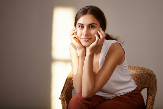 Immagine isolata di carino misterioso giovane femmina in bianco top senza maniche trascorrere del tempo al chiuso tenendosi per mano sul viso, con un affascinante sorriso gioioso, seduto in poltrona all'ora d'oro