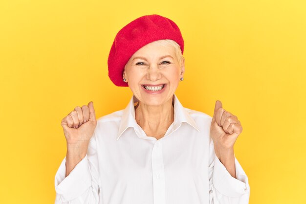 Isolated image of fashionable positive cheerful mature French woman wearing white shirt and red bonnet keeping fists clenched and smiling broadly, overjoyed with good news, screaming Yes excitedly