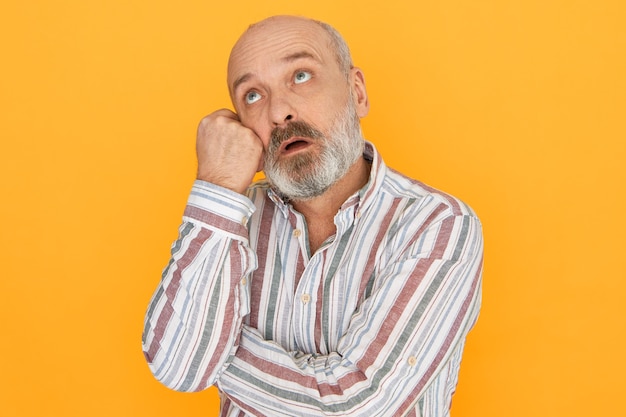 Isolated image of handsome pensive bearded Caucasian male pensioner with puzzled confused expression having memory problems, holding hand on face and looking up, trying to remember something.