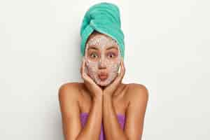 Free photo isolated image of pretty young woman with healthy skin, touches cheeks gently, makes srub facial mask, looks directly, stands bare shoulders