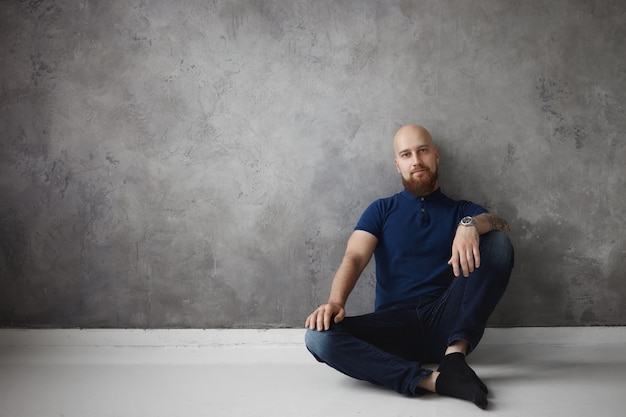 Isolated indoor shot of attractive bald man in stylish polo shirt, jeans and socks relaxing at home, sitting comfortably on floor with hand on his knee, having rest after hard work day at office