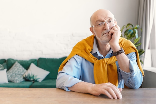 Изолированный снимок привлекательного элегантного шестидесятилетнего пенсионера с белой бородой и лысой головой, сидящего за деревянным столом в гостиной, скучающего, держащего руку за подбородок, задумчивого взгляда
