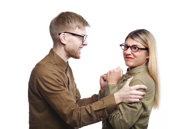 Free photo isolated picture of angry fashionable young couple man and woman wearing shirts and eyeglasses having fight. irritated bearded male shaking his girlfriend by her shoulders