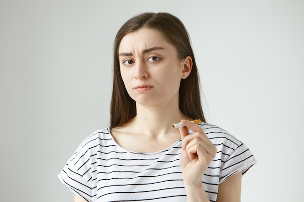 Isolated picture of attractive young brunette female in casual top holding cigarette and frowning, feeling uncertain and doubtful, hesitating to smoke it, trying to break bad habit once and for all