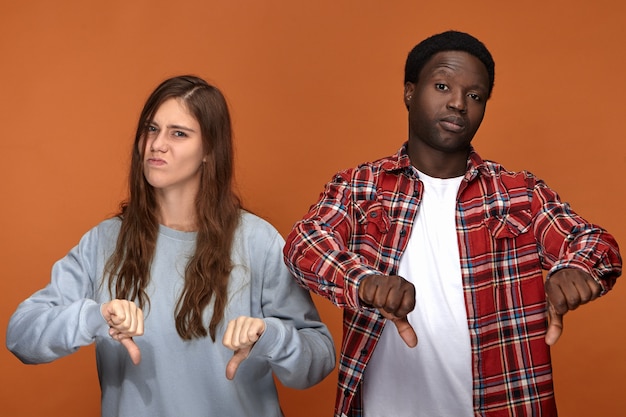 Isolated picture of dissatisfied frowning young European woman and dark skinned man grimacing and making thumbs down gesture, expressing disapproval or dislike, being disappointed with bad movie