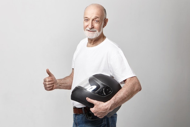オートバイのヘルメットを保持しているカジュアルなTシャツの白いシニア無精ひげを生やしたヨーロッパの男性の孤立した写真