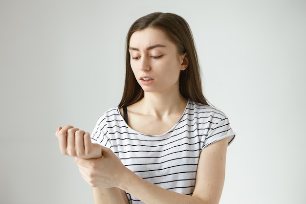 Isolated picture of worried young European woman with dark loose hair holding hand at her wrist while checking pulse preassure after morning workout, having focused serious facial expression
