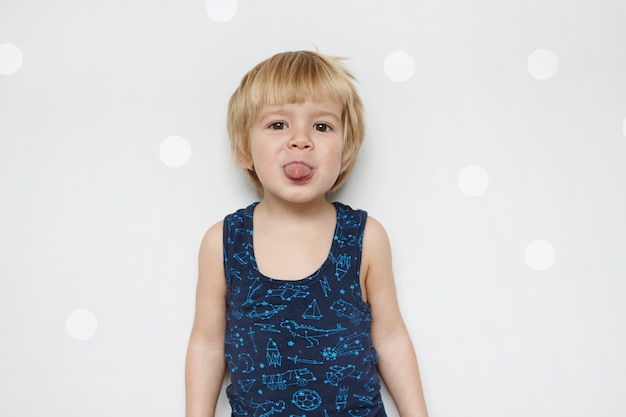 Free photo isolated portrait of adorable funny baby boy with blue eyes wearing tank top having fun indoors, showing his tongue, teasing, standing against wall with copy space for your content