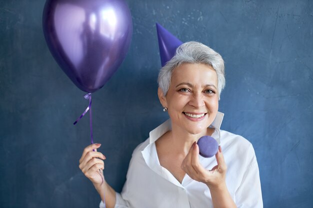 Isolated portrait of cheerful relaxed mature woman in white shirt smiling broadly at camera, holding helium balloon