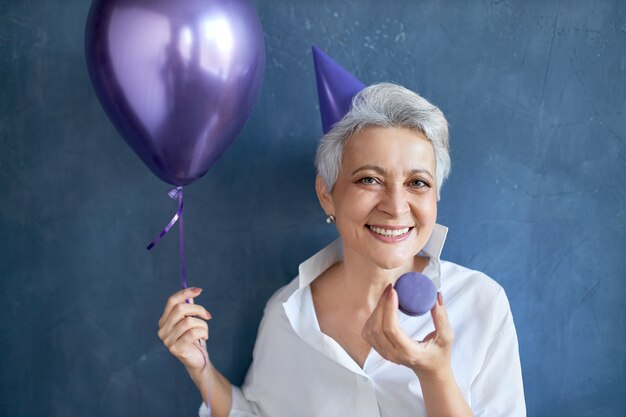 ヘリウム気球を持って、カメラに向かって広く笑っている白いシャツを着た陽気なリラックスした成熟した女性の孤立した肖像画