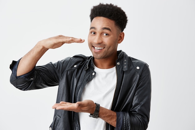 Isolated portrait of cheerful young beautiful black-skinned man with afro hairstyle in white t shirt and leather jacket holding medium size box in hands, looking in camera with happy face expression.