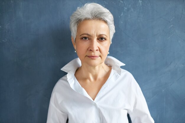 Isolated portrait of confident serious mature female employee with short gray hair frowning posing on blank wall