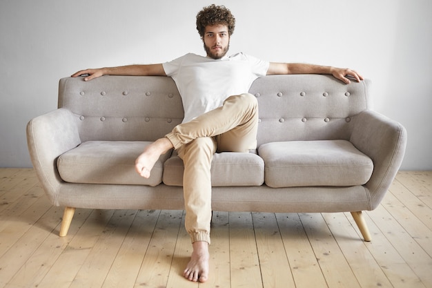 Isolated portrait of fashionable trendy looking young European man with thick beard having rest at home, sitting casually on luxurious sofa, watching TV, feeling relaxed. People, lifestyle and leisure