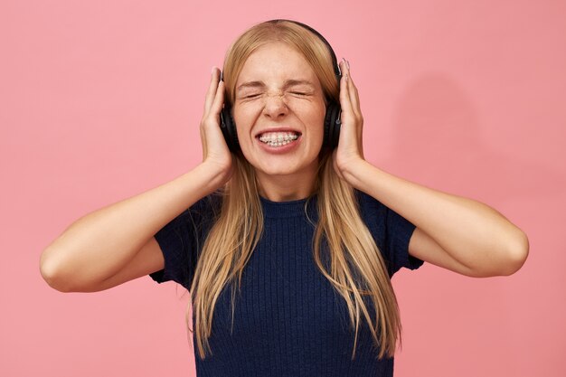 Isolated Portrait of funny emotional young European female with teeth braces wearing wirless headphones closing eyes, holding hands on ears