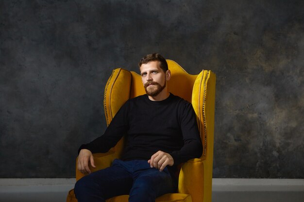 Isolated portrait of good looking elegant young European male with fuzzy trimmed beard and mustache wearing stylish black clothes posing sitting in comfortable yellow armchair alone