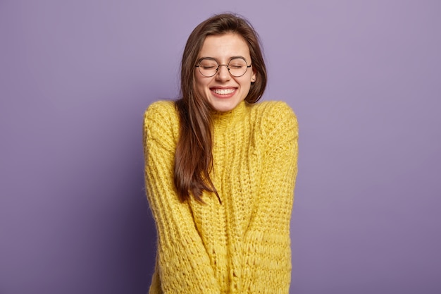 Изолированный портрет счастливой женщины имеет зубастую улыбку, закрывает глаза, испытывает удовольствие от хорошего комплимента, носит очки и желтый джемпер, стоит над фиолетовой стеной. концепция положительных эмоций и чувств