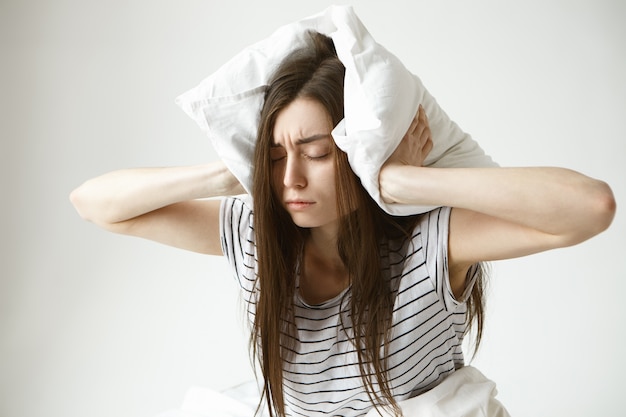 Free photo isolated portrait of stressed young brunette woman wearing striped pajamas covering ears with white pillow feeling frustrated as she can't fall asleep at night because of her snoring husband