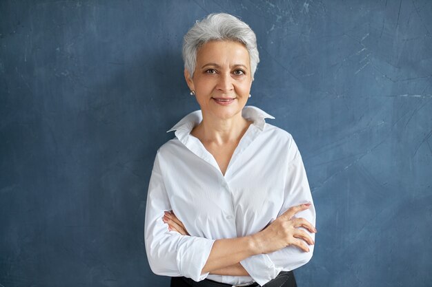 Изолированный портрет стильной успешной 50-летней женщины-брокера в белой рубашке, позирующей на пустой стене