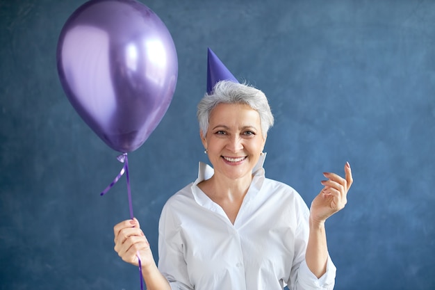 Isolated portrait of successful mature businesswoman with short gray hair posing at blank wall with helium balloon