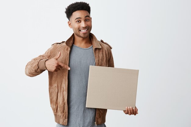 Isolated portrait of young black-skinned man with afro hairstyle in fashionable casual look holding carton board, pointing at it with hand, looking in camera with excited expression