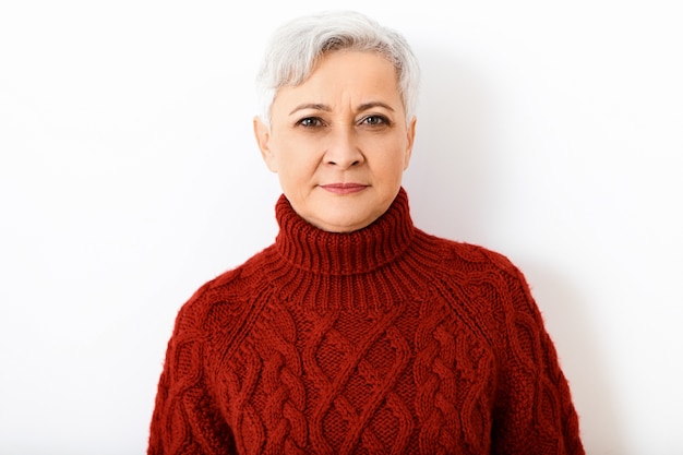 Isolated  serious gray haired retired mature woman in knitted turtleneck sweater having strict concentrated or suspicious facial expression, posing against blank white wall 