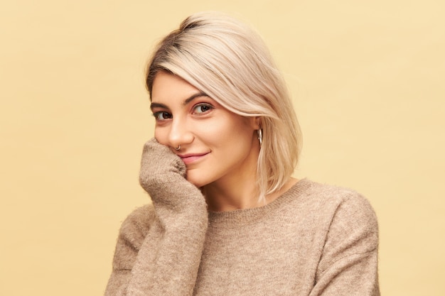 Isolated shot of adorable young European female with dyed bob hairstyle  with curious joyful smile, holding hand under chin, listening attentively. People and lifestyle concept