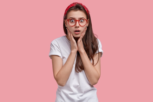 Isolated shot of amazed beautiful lady stupefied by shocking accident, touches face, stands in stupor, wears red headband, white long t shirt