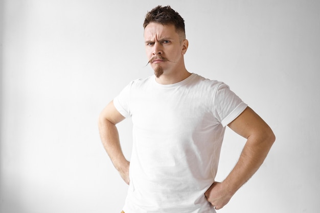 Free photo isolated shot of angry gloomy stylish young european male win stubble and mustache frowning eyebrows and keeping hands on his waist, expressing anger, being mad at his misbehaving children