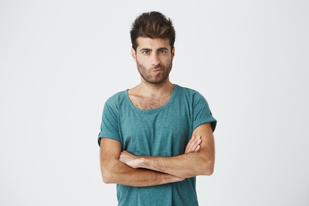 Isolated shot of angry man wearing blue t-shirt with stylish hairstyle holding arms crossed, having skeptical and dissatisfied look. Distrust, skepticism and doubt