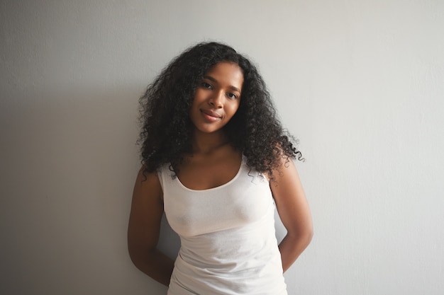 Isolated shot of attractive charming young African American woman with voluminous black hair and clean perfect skin posing at blank wall in white tank top, with shy cute expression