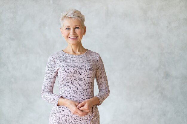 Isolated shot of attractive confident European woman with short haircut posing against textured marbled wall, wearing elegant pencil dress, holding hands together and smiling happily