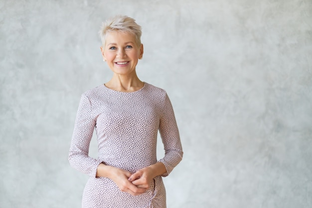 Isolated shot of attractive confident European woman with short haircut posing against textured marbled wall, wearing elegant pencil dress, holding hands together and smiling happily