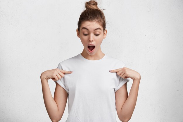 Isolated shot of attractive female with amazed look, keeps mouth widely opened, indicates at blank t shirt, poses against white studio background, expresses surprisment. People, clothing, advertisment