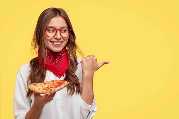 Изолированный снимок привлекательной улыбающейся девушки показывает направление в пиццерию, ест вкусную пиццу с сыром и помидорами, указывает большим пальцем на место для копирования у желтой стены. Женщина перекусывает в помещении