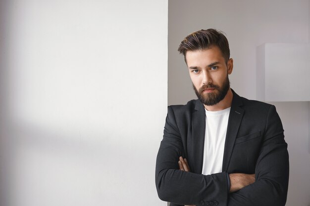 Isolated shot of attractive successful young brunette bearded male entrepreneur wearing trendy jacket over casual white t-shirt keeping his arms folded, expressing reluctancy or disagreement