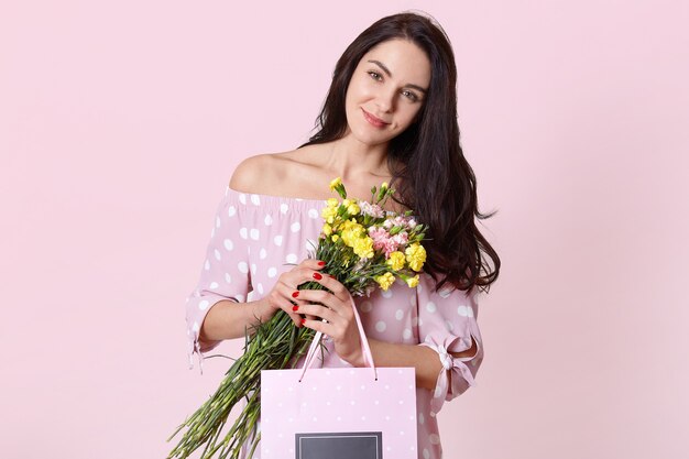 Isolated shot of attractive young European woman has black long hair, wears polka dot dress, holds gift bag and flowers, poses on light pink wall, celebrates International Womens day