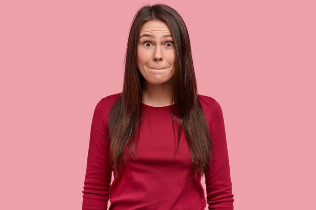 Isolated shot of attractive young woman with long black hair, presses lips, has surprised expression, wears causal red clothes, poses over pink background