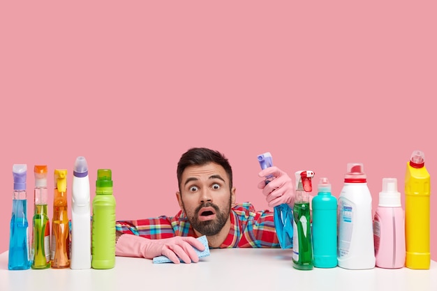 Isolated shot of bearded man has shocked expression, stares at you, holds spray, uses rag for cleaning