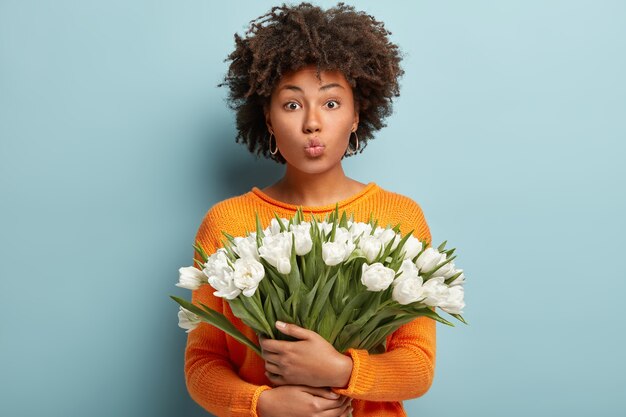 Isolated shot of beautiful black young woman with curly hairstyle, holds nice bouquet of white tulips, keeps lips folded, wears orange jumper, isolated over blue wall. Spring time concept