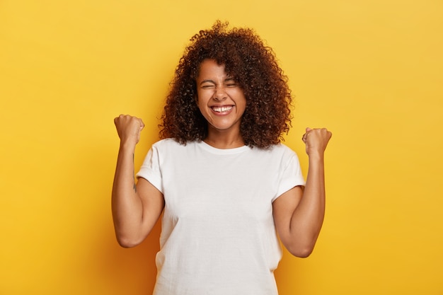 Isolated shot of beautiful successful woman with curly hair, raises clenched fists, celebrates triumph, being very pleased and happy, closes eyes from pleasure, wears white t shirt. Yeah, I did it!