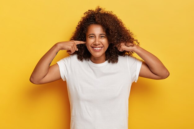 Isolated shot of calm happy woman plugs ears, has fun at party, ignores loud music, feels relaxed and pleased, hears no noise, has funny expression, dressed in white wear, models over yellow wall