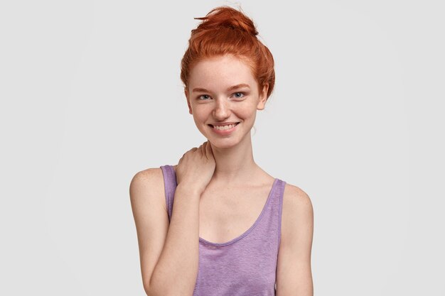 Isolated shot of carefree cheerful freckled red haired young woman keeps hand on neck, has friendly expression, healthy skin, wears casual vest, models against white wall. Beauty concept