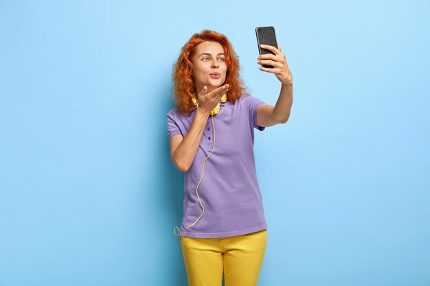 Isolated shot of charming lovely curly red haired cheerful woman sends air kiss at camera of cell phone