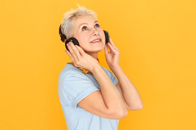 Isolated shot of charming thoughtful middle aged European woman using wireless headset listening to classic music tracks enjoying high quality mp3 sound, looking up with dreamy facial expression