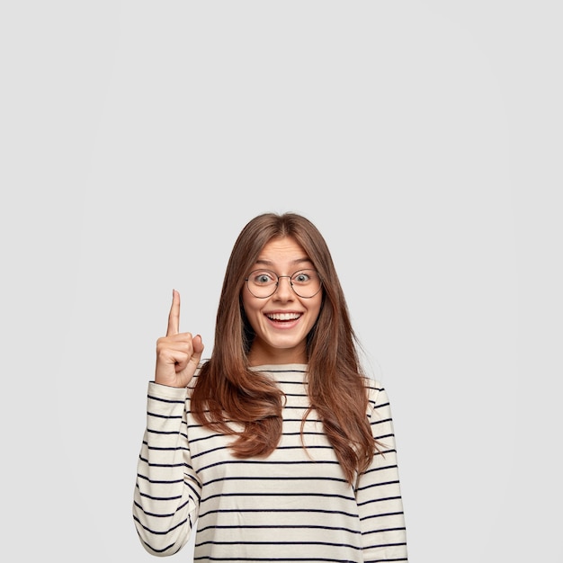 Изолированный снимок веселой кавказской женщины с прямыми волосами, показывает свободное пространство, показывает указательным пальцем для вашего рекламного контента, носит повседневный полосатый свитер, изолированный на белой стене